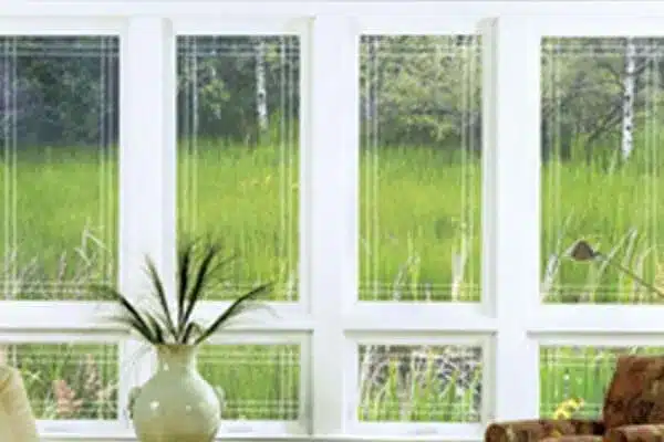 View from inside a room showing large white-framed windows opening to a lush green landscape with tall grass and trees. A light-colored vase with decorative grass sits on a table in the foreground.