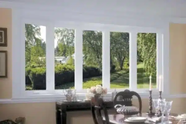 A dining room window with white frames offers a view of a lush green garden. Inside, a table is elegantly set with candles, glassware, and a floral centerpiece, creating a serene and inviting ambiance.