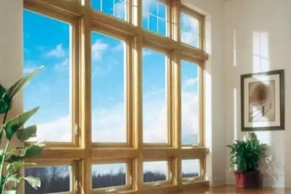Large floor-to-ceiling windows with wooden frames showing a clear blue sky and clouds. Sunlight streams in, illuminating a potted plant and picture frame on the wall, creating a bright and inviting atmosphere.