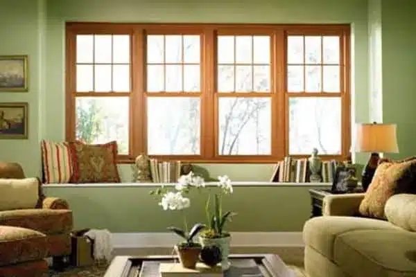 Cozy living room with green walls, a beige sofa, and an armchair. Large wooden-framed windows in the background let in natural light. A coffee table with potted orchids is in the center, and books are displayed on the windowsill.