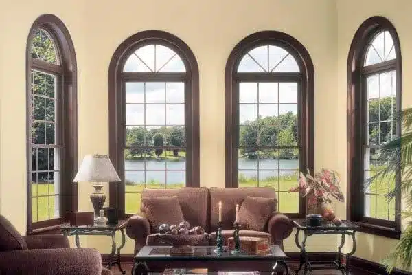 A cozy living room with large arched windows overlooking a serene lake and green landscape. The room features a brown sofa, a lamp, a candle, and a plant on wooden side tables. Soft natural light fills the space, creating a warm ambiance.