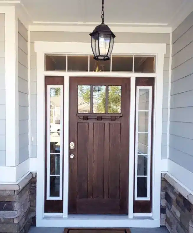 A wooden front door with a rectangular window at the top, surrounded by narrow glass panels on each side. The door is set in a light-colored frame, with a hanging lantern above providing light. The entrance is bordered by light gray siding and stone accents.