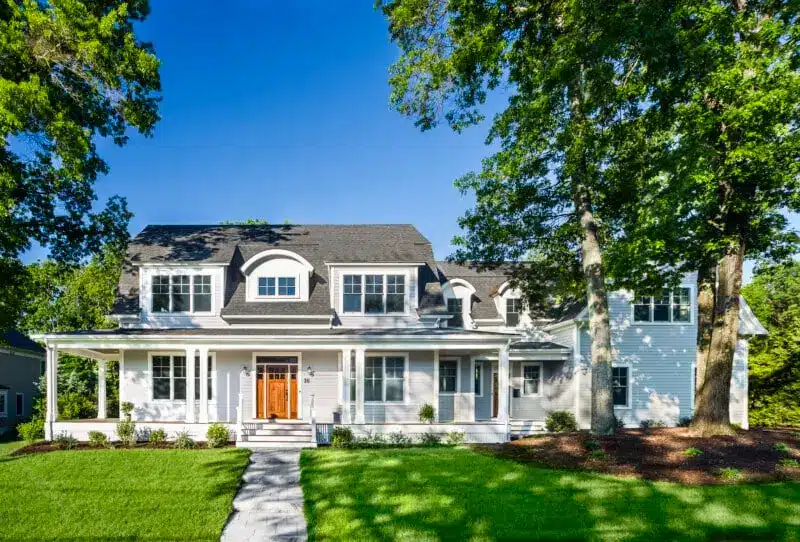 A two-story white house with a dark roof, featuring large windows and a wooden front door. It has a wraparound porch and is surrounded by green grass and tall trees under a clear blue sky. A stone pathway leads to the entrance.
