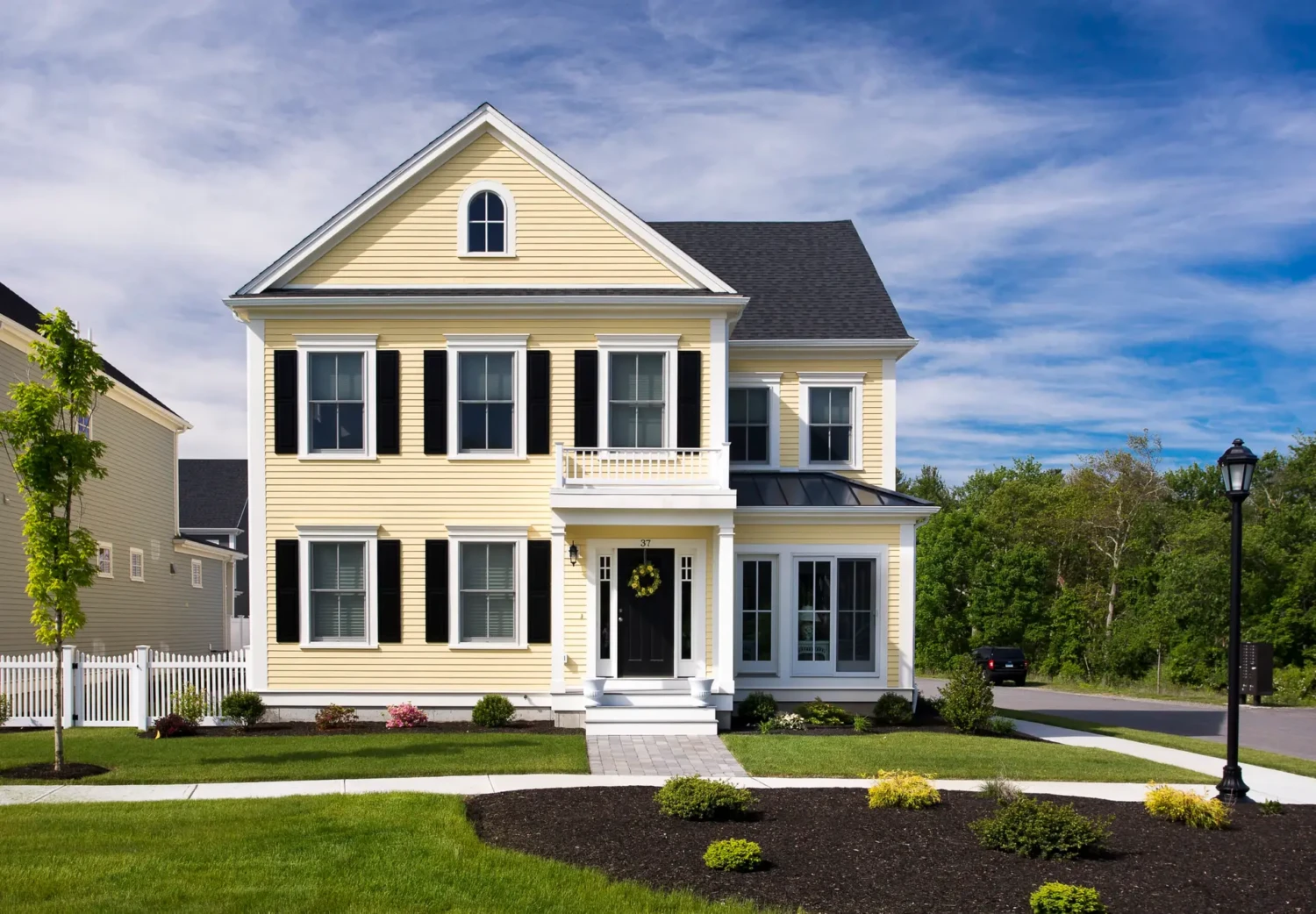 A two-story, yellow house with black shutters and a small balcony over the front porch. It features a neatly maintained garden and a driveway. The sky is blue with light clouds, and a lamppost stands on the right side near the driveway.