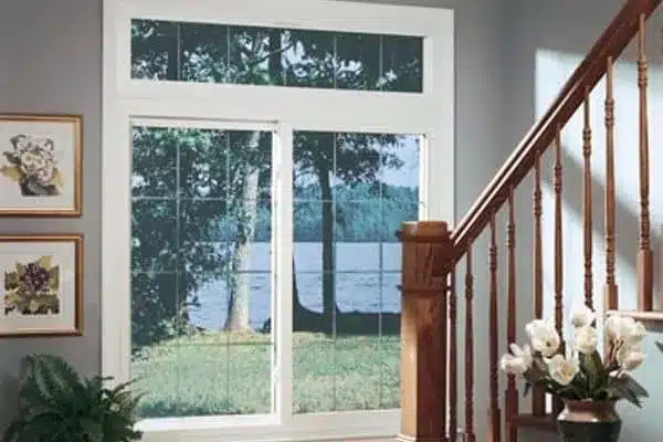 A staircase with wooden railings is on the right. To the left, a large window displays a scenic view of trees and a lake. Two framed floral paintings and a potted plant are on the gray walls. White flowers are placed on a table.