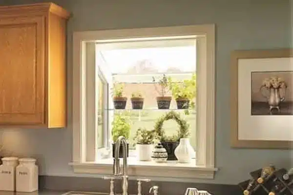 A kitchen window display featuring potted plants on shelves, with sunlight streaming through. The countertop has a silver faucet and beige canisters. On the right, a framed black-and-white photo of flowers hangs on the wall.
