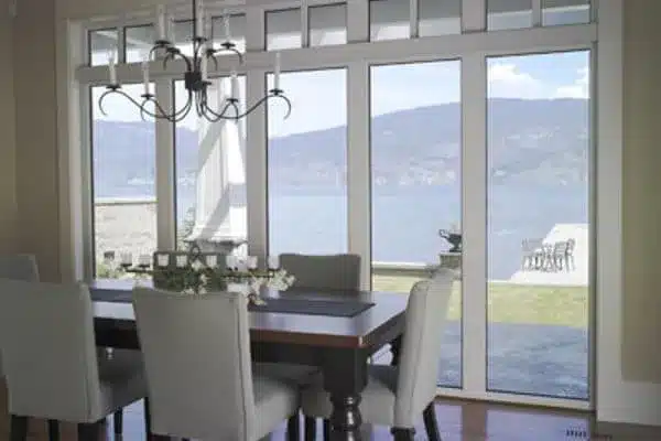 Dining room with a wooden table and upholstered chairs. A chandelier hangs above. Large glass doors reveal a scenic lake view with mountains in the background and an outdoor patio.
