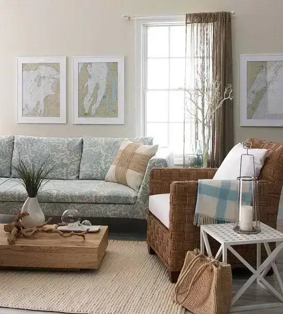 A cozy living room with a patterned sofa, a wicker armchair, and a wooden coffee table. Three framed maps hang on the wall. A beige rug covers the floor, and a window with sheer curtains brightens the space. Accessories include plants and a lantern.
